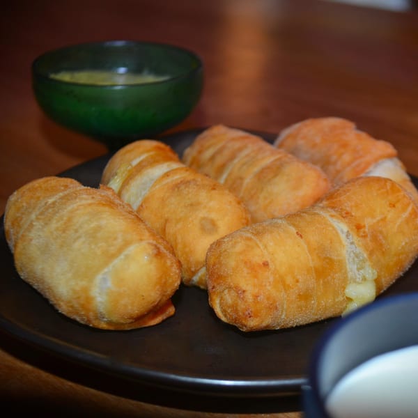 Golden-brown Venezuelan tequeños, cheese oozing, on a dark plate with a green dipping sauce bowl.