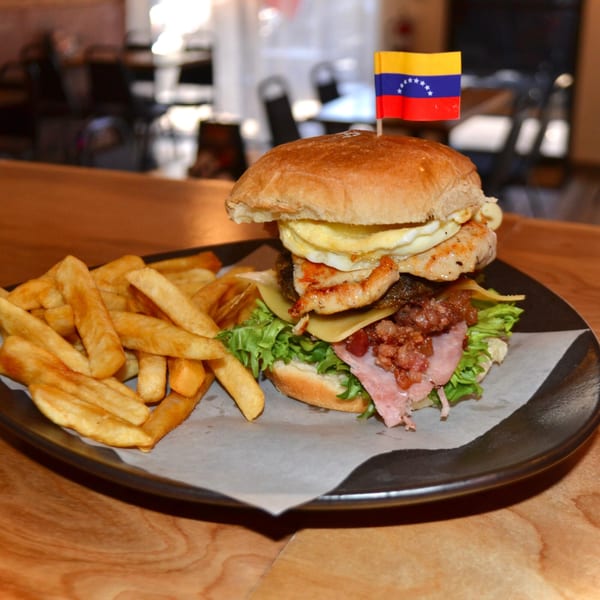 A Venezuelan burger with a flag, chicken, ham, cheese, and fries on a dark plate.