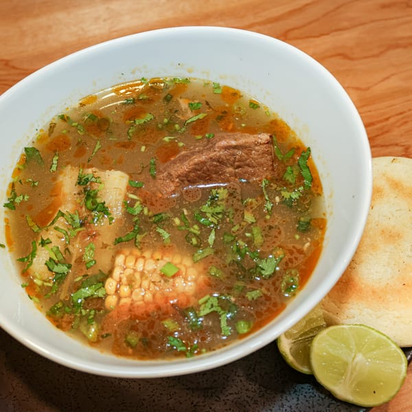 Venezuelan beef soup with corn, cilantro, and yuca, served with arepas and lime wedges.