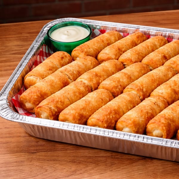 A foil tray of golden-brown Venezuelan tequeños with a side of white dipping sauce.